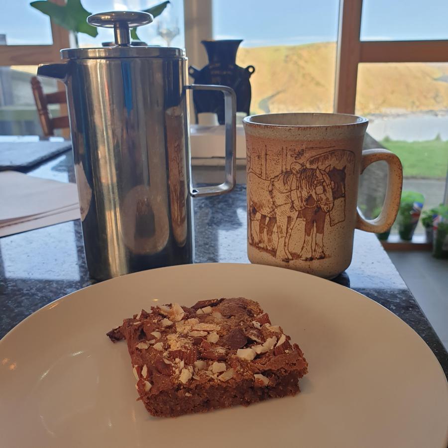 Chickpea blondies work well with coffee. This shows a slice on a plate with a brown coffee mug and siler cafetiere behind.